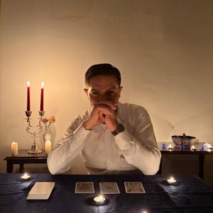 Puede incluir: Una persona con una camisa blanca est&aacute; sentada en una mesa con cartas del tarot y velas encendidas. Dos velas rojas altas est&aacute;n en un candelabro plateado. Otras velas est&aacute;n esparcidas por la mesa.