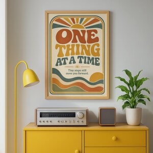 May include: A framed wall art print featuring a sunburst graphic and wavy lines in orange, yellow, and teal. The text reads "ONE THING AT A TIME" in large, bold letters, followed by the smaller text "Tiny steps still move you forward."