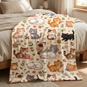May include: A cream-colored throw blanket featuring a repeating pattern of various cartoon cats in shades of orange, gray, black, and white, interspersed with colorful flowers, small hearts, paw prints, and balls of yarn.