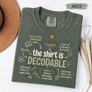 Op de afbeelding: Een mosgroen t-shirt met een grafische print met de tekst "the shirt is DECODABLE" omringd door educatieve taalkundige notities zoals "Closed Syllable", "Digraphs", "R-controlled" en "Silent-e" met diverse kleine iconen.
