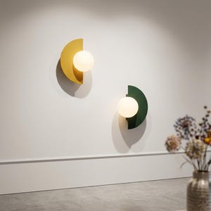 Puede incluir: Dos apliques de pared modernos. Uno es un semicírculo amarillo mostaza, el otro verde oscuro, cada uno con una bombilla esférica y brillante. Los accesorios están montados en una pared blanca, ofreciendo un diseño minimalista.