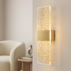 Puede incluir: Aplique de pared moderno con un panel rectangular de acrílico transparente con inclusiones de pan de oro. Una banda horizontal de metal dorado cepillado y un soporte de pared a juego completan el diseño. El aplique está montado en una pared neutra.