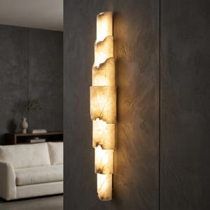 Puede incluir: Aplique de pared vertical compuesto por bloques rectangulares de alabastro apilados. El alabastro, de color blanco roto, está iluminado y presenta vetas naturales y bordes irregulares. El aplique está fijado a una pared gris oscuro, creando contraste.