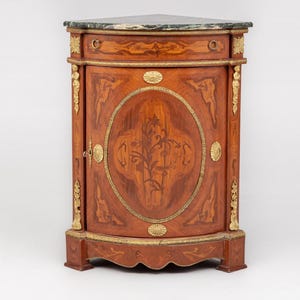 May include: A corner cabinet with a marble top and a curved front. The cabinet is made of wood with a reddish-brown finish and gold-colored accents. The door features an oval inlay with a floral design.