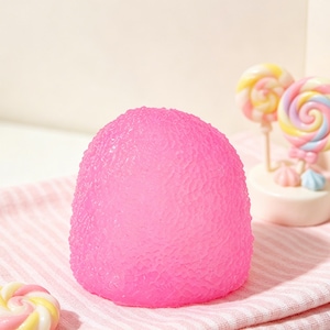 Op de afbeelding: Een felroze, getextureerd, koepelvormig object ligt op een roze en wit gestreepte doek. Twee kleurrijke lolly's op stokjes staan op de achtergrond. De afbeelding heeft een zachte, pastelkleurige uitstraling.