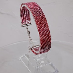 May include: A beaded bracelet featuring a vibrant pink and red gradient pattern. The bracelet is constructed with small, uniform beads in a structured, curved band shape, finished with silver-toned metal end caps and a delicate silver chain extender.