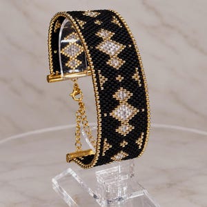 Puede incluir: Una pulsera ancha tipo brazalete hecha de cuentas de vidrio negras, doradas y transparentes con un patrón geométrico de diamantes. La pulsera cuenta con terminales de metal dorado y una cadena extensora dorada con cierre de mosquetón.