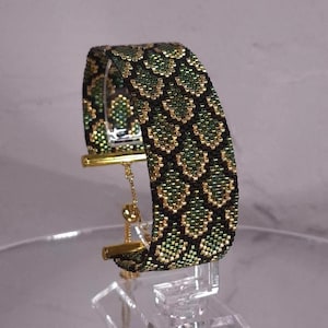 Puede incluir: Una pulsera ancha de cuentas tejidas con un patrón de escamas repetitivo en oro metálico, verde bosque y negro. La pulsera incluye un mecanismo de cierre deslizante en tono dorado con una cadena fina para un ajuste regulable en la muñeca.