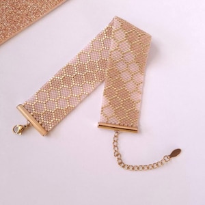 May include: A wide choker necklace crafted from small, woven beads in a repeating honeycomb pattern of soft pink and metallic gold. The piece features gold-toned metal end bars, a lobster claw clasp, and an adjustable chain with a small oval charm.