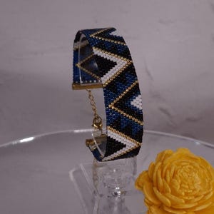 May include: A beaded bracelet featuring a geometric chevron pattern in navy blue, black, and white, accented with thin lines of metallic gold beads. The bracelet includes a gold-toned metal clasp and an adjustable chain link extension.
