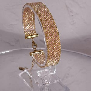 Può includere: Un braccialetto di perline intrecciate con un motivo ripetuto di perline color oro metallizzato e rosa tenue. Il braccialetto è rifinito con terminali in metallo color oro, una chiusura a moschettone e una catenella di estensione regolabile.