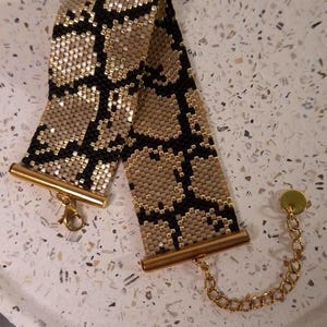 May include: A wide beaded bracelet featuring a repeating black and metallic gold snake skin pattern. The bracelet is finished with gold-toned metal end bars, a lobster claw clasp, and a short gold chain extender with a small circular charm.