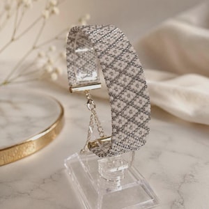 May include: A woven beaded bracelet featuring a repeating diamond pattern in white and metallic silver. The bracelet includes silver-toned metal end caps and a delicate silver chain extender with a lobster claw clasp, displayed on a clear stand.