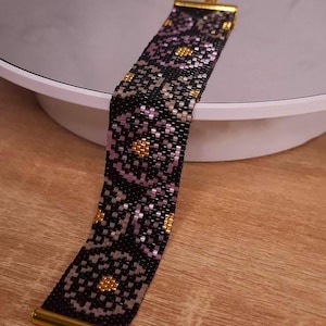 May include: A wide beaded bracelet featuring a repeating floral pattern in black, metallic gold, and soft lavender beads. The bracelet is finished with polished gold-tone metal end bars and a delicate gold-tone chain extension at one end.