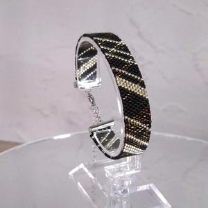 Op de afbeelding: Een geweven kralenarmband met een herhalend diagonaal patroon van zwarte, metallic bronskleurige en zilveren kralen. De armband heeft een zilverkleurige metalen sluiting en een verstelbare ketting, gepresenteerd op een doorzichtige acrylaat standaard.