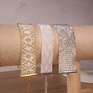 May include: Three beaded bracelets featuring a flat, wide design. The left bracelet has a pink and gold geometric pattern. The middle bracelet is solid iridescent white. The right bracelet is solid metallic silver. Each has a metal end bar and chain.