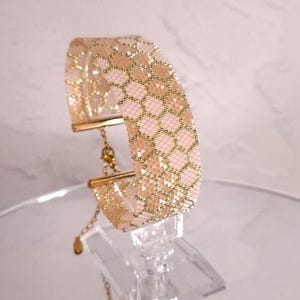 Op de afbeelding: Een brede kralenarmband met een herhalend honingraatpatroon in zacht poederroze en metallic goud. De armband is afgewerkt met goudkleurige metalen eindstukken en een fijne verstelbare ketting met een kleine goudkleurige bedel aan het uiteinde.