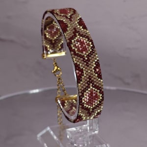 May include: A beaded bracelet featuring a geometric pattern in shades of deep red, light tan, and metallic gold. The bracelet is constructed from small, uniform beads and includes a gold-toned metal chain extender with a lobster claw clasp.