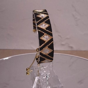 May include: A beaded bracelet featuring a geometric pattern of black, white, and gold beads. The bracelet has a structured, curved band design with a gold-toned adjustable sliding chain closure with small spherical ends for a secure fit.