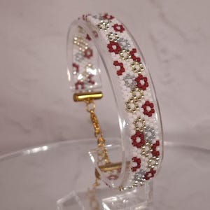 May include: A beaded bracelet featuring a repeating pattern of white, deep red, and metallic silver seed beads. The band is connected to a gold-toned metal clasp and an adjustable chain link extension, creating a structured and colorful accessory.