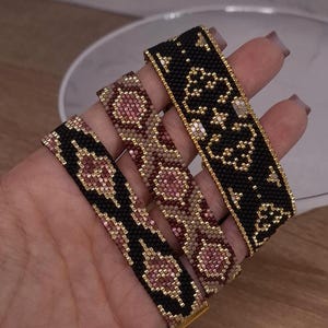 May include: Three flat woven bead bracelets featuring geometric patterns. Two bracelets are primarily black with pink and gold accents, while the centre bracelet features a repeating pattern of pink, gold, and tan beads. Each bracelet has gold metal ends.