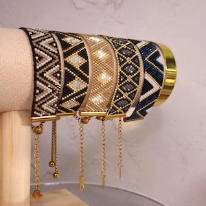 Könnte beinhalten: Fünf Perlenarmbänder mit geometrischen Mustern in Schwarz, Weiß, Gold, Grau und Blau. Jedes Armband hat einen goldfarbenen Metallverschluss und eine feine Goldkettenverlängerung mit einem kleinen Anhänger, nebeneinander auf einem Ständer.