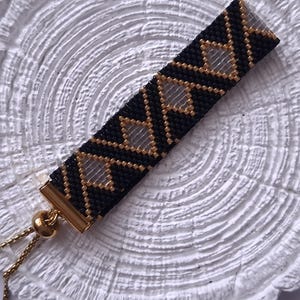 May include: A rectangular beaded bracelet featuring a geometric pattern of black and metallic gold seed beads with clear diamond-shaped cutouts. The bracelet includes a gold-toned metal slider clasp with thin gold chains for an adjustable fit.