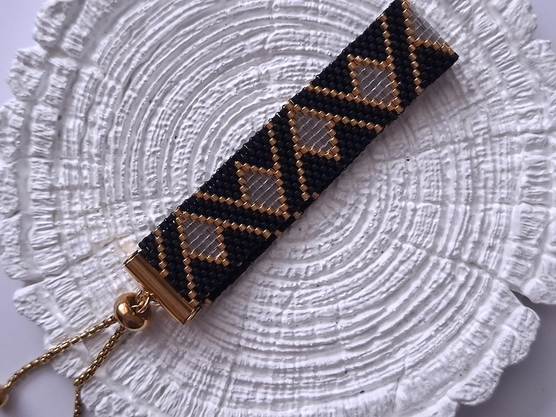 May include: A rectangular beaded bracelet featuring a geometric pattern of black and metallic gold seed beads with clear diamond-shaped cutouts. The bracelet includes a gold-toned metal slider clasp with thin gold chains for an adjustable fit.