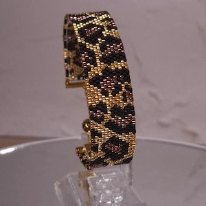 Op de afbeelding: Een manchetarmband gemaakt van kleine glaskralen in een herhalend luipaardpatroon. Het ontwerp heeft een kleurenpalet van metallic goud, diepzwart en bronsbruine kralen, wat zorgt voor een gestructureerd en getextureerd oppervlak.