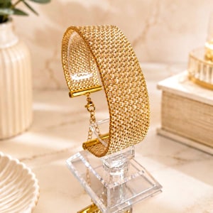 Può includere: Un bracciale in maglia color oro caratterizzato da una trama metallica finemente intrecciata. Il bracciale presenta una chiusura rettangolare su ciascuna estremità e include una delicata catenella di estensione dorata con moschettone.