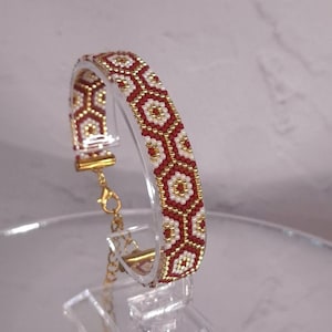 May include: A beaded bracelet featuring a geometric pattern of red, white, and metallic gold seed beads. The bracelet includes a gold-toned metal clasp and an adjustable chain link extension, showcasing a repeating hexagonal design throughout the band.