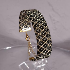 May include: A woven bead bracelet featuring a repeating geometric diamond pattern in black and metallic gold. The bracelet includes gold-toned metal end caps, a lobster claw clasp, and an adjustable chain link extension for a secure fit.
