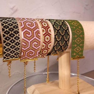 May include: Five wide beaded bracelets featuring intricate geometric patterns in black, gold, white, pink, red, and green. Each bracelet has a metallic gold-tone clasp and a delicate gold-tone chain extender hanging from the bottom edge.