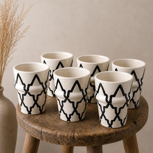 Könnte beinhalten: Sechs weiße Keramikbecher mit einem schwarzen geometrischen Muster stehen auf einem kleinen, runden Holzschemel. Die Becher haben einen ausgestellten oberen Rand und ein dekoratives Band am Boden. Im Hintergrund eine Vase mit getrocknetem Gras.