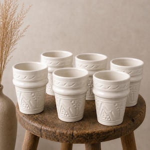Peut inclure: Six tasses en céramique blanc cassé avec des motifs floraux et géométriques en relief sont disposées sur un petit tabouret rond en bois. Les tasses ont un design texturé et détaillé et un bord légèrement évasé. Un vase avec des plantes séchées est en arrière-plan.