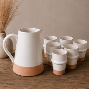 Peut inclure: Un pichet en céramique blanche avec une base et une anse couleur terre cuite, accompagné de six tasses en céramique blanche assorties avec des bases couleur terre cuite. L'ensemble est disposé sur une surface en bois rustique, avec un vase de plantes séchées en arrière-plan.