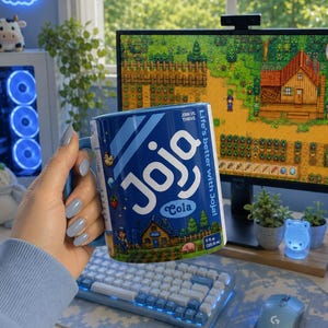 Könnte beinhalten: Eine blaue Keramik-Tasse mit dem Text "Joja Cola" und einer Cartoon-Illustration. Die Tasse wird vor einem Computermonitor gehalten, der eine Videospielszenen anzeigt. Die Tasse ist 325 ml.