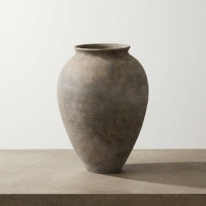Könnte beinhalten: Eine große, neutral gefärbte Vase mit breitem Körper und schmalem Hals. Die Vase hat eine strukturierte Oberfläche und ein leicht verwittertes Aussehen. Die Vase steht auf einer Betonoberfläche. Die Gesamtästhetik ist rustikal und minimalistisch.