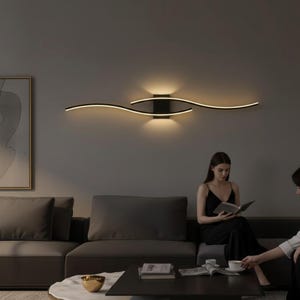 Op de afbeelding: Een moderne, zwarte wandlamp met een gebogen, golfachtig ontwerp. De lamp straalt een warme, uitnodigende gloed uit en verlicht de ruimte boven een donkergrijze bank. De kamer heeft een minimalistische esthetiek.