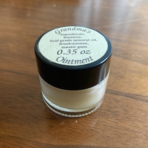 May include: A small glass jar of ointment with a black lid. The label reads "Grandma's" and lists ingredients like beeswax and frankincense. The jar contains 0.35 oz of ointment.