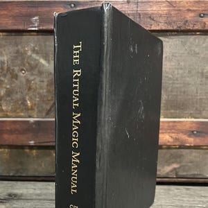 May include: A black book titled "The Ritual Magic Manual" by David Griffin. The title and author's name are in gold lettering. The book is standing upright against a wooden background.