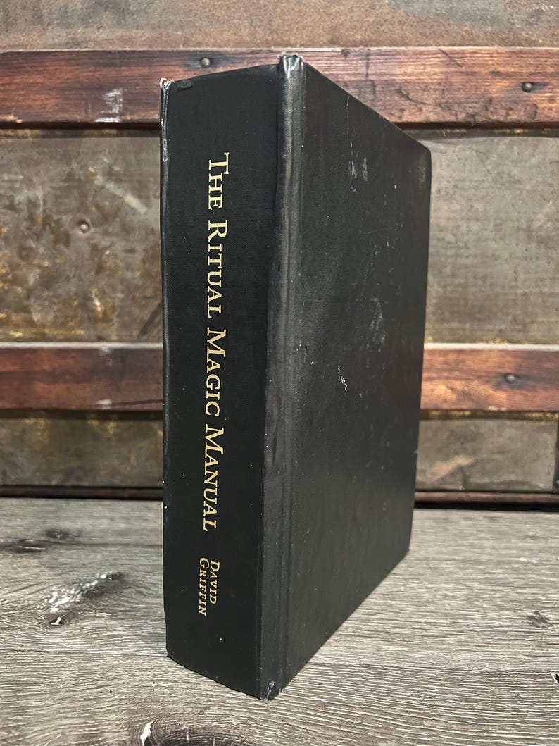 May include: A black book titled "The Ritual Magic Manual" by David Griffin. The title and author's name are in gold lettering. The book is standing upright against a wooden background.