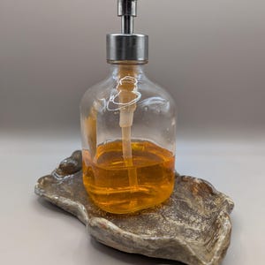 Può includere: Un dispenser di sapone in vetro trasparente con una pompa argentata, riempito di liquido arancione, poggia su un portasapone in ceramica grigia strutturata. Il dispenser ha una forma arrotondata e un lungo beccuccio. Il portasapone ha una forma irregolare.