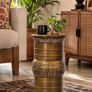 Könnte beinhalten: Ein dekorativer Beistelltisch aus Messing mit einer runden Holzplatte. Der Tisch hat eine zylindrische Form mit aufwendigen Ausschnittmustern und horizontalen Bändern. Eine schwarze Tasse und eine kleine Topfpflanze stehen darauf. Der Tisch steht auf einem gemusterten Teppich. Höhe ca. 60 cm.