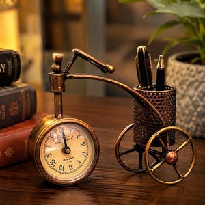 Könnte beinhalten: Ein dekorativer Schreibtisch-Organizer in Form eines Fahrrads, mit einer Uhr und einem Stifthalter. Das Zifferblatt zeigt römische Ziffern und den Text "Village Clock Birmingham est 1790". Der Stifthalter ist ein kleiner, perforierter Metallkorb.