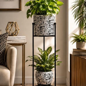 Puede incluir: Un soporte de metal negro para plantas con dos macetas con plantas verdes. Las macetas tienen un fondo blanco con letras negras. El soporte está en una habitación luminosa con una paleta de colores neutros.