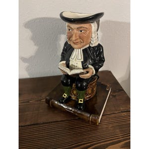 Może przedstawiać: Ceramiczna figurka mężczyzny w czarnym płaszczu i kapeluszu, siedzącego i czytającego książkę. Figurka stoi na brązowej podstawie w kształcie książki. Mężczyzna ma białe włosy i biały kołnierz. Przedmiot dekoracyjny.