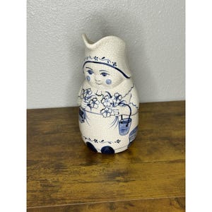 Może przedstawiać: Ceramiczny dzban w kształcie kobiety, pomalowany na biało i niebiesko. Dzbanek ma wykończenie typu craquelé i przedstawia kobietę trzymającą kwiaty i wiadro. Projekt zawiera motywy kwiatowe i domowe.