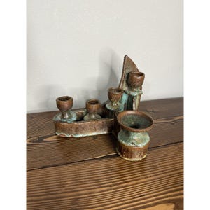May include: A handcrafted ceramic candle holder with a brown and turquoise glaze. The piece features four candle cups and a decorative, abstract design. The candle holder is placed on a wooden surface.