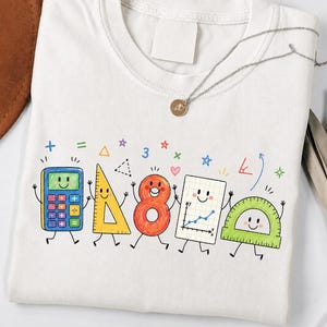 May include: A white crew neck t-shirt featuring a colourful graphic print of anthropomorphic school supplies including a blue calculator, yellow triangle, orange number eight, graph paper, and a green protractor with smiling faces.